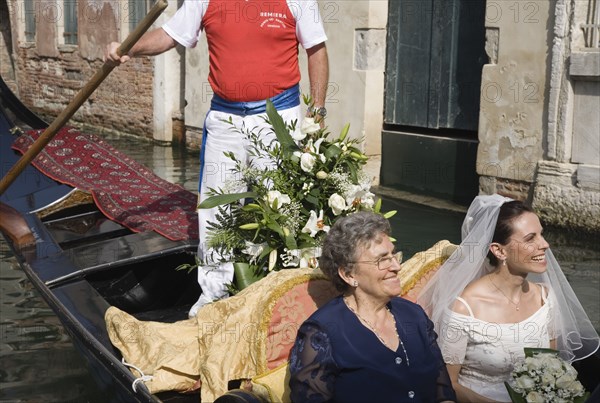 Venice, Veneto, Italy. Cropped shot of gondolier steering decorated gondola with smiling bride in wedding dress and veil carrying bouquet and her mother on wedding trip on canal in late summer sunshine. Italy Italia Italian Venice Veneto Venezia Europe European City Wed Wedding Marriage Dress Bride White Lace Veil Canal Gondola Gondolier Smiling Happy Flowers Bouquet Contented Marriage Marrying Espousing Hymeneals Nuptials Mum Religion Southern Europe