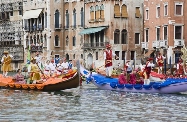 Venice, Veneto, Italy. Participants in the Regata Storico historical regatta held annually in September wearing traditional costume approaching the Rialto bridge in brightly painted gondolas with onlookers gathered on the balconies of canalside buildings behind. Teams represent Sestiere districts of Venice in traditional races. Italy Italia Italian Venice Veneto Venezia Europe European City Regata Regatta Gondola Gondola Gondolas Gondolier Boat Architecture Exterior Water Classic Classical Destination Destinations History Historic Holidaymakers Older Southern Europe Tourism Tourist