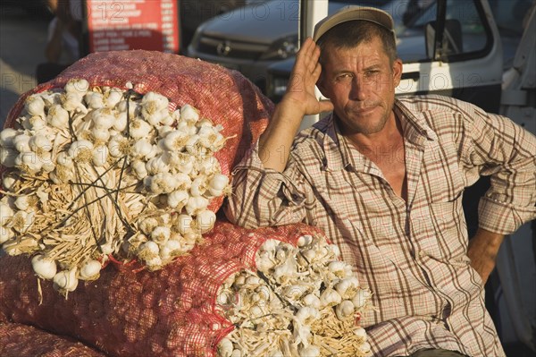 Kusadasi, Aydin Province, Turkey. Stallholder at weekly market leaning on sacks of garlic in the late afternoon summer sunshine. Turkey Turkish Eurasia Eurasian Europe Asia Turkiye Aydin Province Kusadasi Market markets Stall Vendor Man Male Garlic Sacks Destination Destinations European Male Men Guy Middle East One individual Solo Lone Solitary South Eastern Europe Western Asia