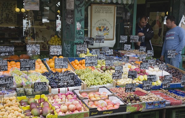 Vienna, Austria. The Naschmarkt. Owners of market fruit stall standing behind shopfront display that includes mangoes grapes pomegranates mangosteens and apricots. Austria Austrian Republic Vienna Viennese Wien Europe European City Capital Naschmarkt Market Display Fresh Food Fruit Vegetables Veg Vegetable Stall Shop Store Destination Destinations One individual Solo Lone Solitary Osterreich Viena Western Europe