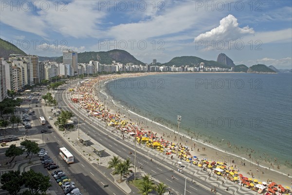 Rio de Janeiro, Brazil. Copacabana. The full sweep of Avenue Atlantica mosiac sidewalk with crowds on the beach and in the sea. Umbrellas hotels tour buses Sugarloaf Mountain blue sea and sky with puffy cloud. American Brasil Brazilian Destination Destinations Latin America Latino Sand Sandy Beaches Tourism Seaside Shore Tourist Tourists Vacation South America Southern Sunbather Travel Water