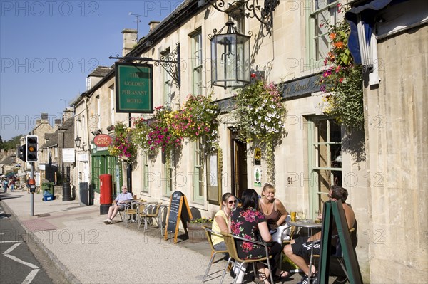 Burford, Gloucestershire, England. Tourists sat outside the Golden Pheasant Hotel in the Cotswolds. England English UK United Kingdom GB Great Britain British Gloucestershire Cotswold Cotswolds Burford High Street Golden Pheasant Hotel Tourist Tourists Sat Outside Tables Blue British Isles Destination Destinations European Great Britain Holidaymakers Northern Europe Sightseeing Tourism United Kingdom