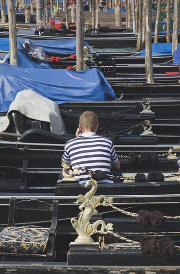 Venice, Veneto, Italy. Gondolier in striped shirt using mobile phone while sitting in one of a line of moored gondolas with back to camera. Italy Italia Italian Venice Veneto Venezia Europe European City Gondola Gondolas Gondolier Phone Mobile Cell Telephone Stripe Srripes Striped Shirt Water 1 Cellular Color Destination Destinations One individual Solo Lone Solitary Single unitary Southern Europe