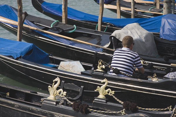 Venice, Veneto, Italy. Gondolier wearing striped shirt using mobile phone sitting in one of a line of moored gondolas with back to camera. Italy Italia Italian Venice Veneto Venezia Europe European City Water Shirt Striped Stripes Stripe Stripe Cell Mobile Phone Gondolier Gondolas Gondola 1 Cellular One individual Solo Lone Solitary Single unitary Southern Europe