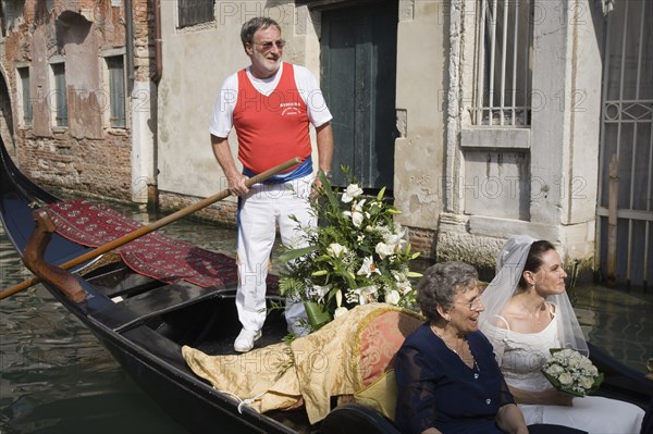 Venice, Veneto, Italy. Gondolier steering decorated gondola carring bride in wedding dress and veil holding bouquet and her mother on wedding trip on canal in late summer sunshine. Italia Italian Venice Veneto Venezia Europe European City Wed Wedding Marriage Dress Bride White Lace Veil Canal Gondola Gondolier Smiling Happy Flowers Bouquet Contented Marriage Marrying Espousing Hymeneals Nuptials Mum Religion Southern Europe