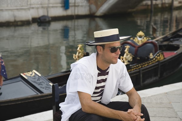 Venice, Veneto, Italy. Centro Storico Young gondolier in traditional uniform of striped shirt and straw hat with black sash wearing sunglasses seated beside moored gondola on canal in late summer sunshine. Italy Italia Italian Venice Veneto Venezia Europe European City Centro Storico Gondola Gondolas Gondolier People Man Hat Boater Water Canal Sunglasses Shades Sun Classic Classical Destination Destinations Historical Immature Male Men Guy Older One individual Solo Lone Solitary Southern Europe