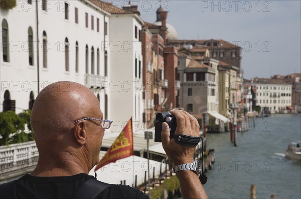 Venice, Veneto, Italy. Male tourist making a video recording from bridge looking along canal in late summer sunshine. Italy Italia Italian Venice Veneto Venezia Europe European City Water Canal Tourist Man Camera Video Recording Bald Glasses Destination Destinations Holidaymakers Male Men Guy One individual Solo Lone Solitary Sightseeing Southern Europe Tourism Tourists