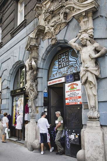 Budapest, Pest County, Hungary. Art Nouveau sculpture on facade of apartment building with local people standing in doorway below and outside shopfront on left. Hungary Hungarian Europe European East Eastern Buda Pest Budapest City Art Nouveau Sculpture Facade Building Architecture Facade Shopfront Aprtment Apartments Flats Entrance Door Doorway People Destination Destinations Eastern Europe Flat