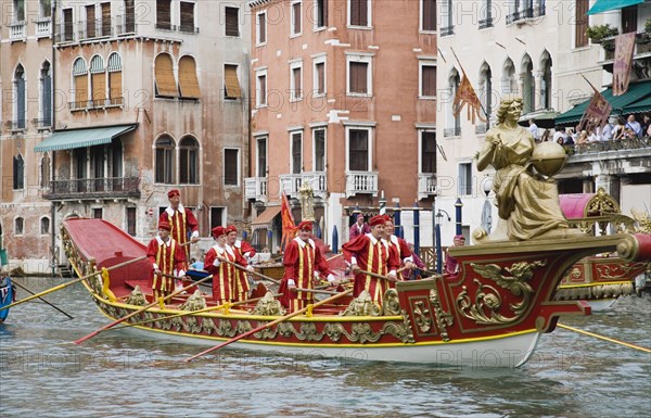 Venice, Veneto, Italy. Participants in the Regata Storico historical regatta held annually in September wearing traditional costume and rowing red and gold painted gondola approaching the Rialto bridge with onlookers gathered on the balconies of canalside buildings. Teams represent Sestiere districts of Venice in traditional races. Italy Italia Italian Venice Veneto Venezia Europe European City Regata Regatta Gondola Gondola Gondolas Gondolier Boat Architecture Exterior Water Classic Classical Destination Destinations History Historic Holidaymakers Older Southern Europe Tourism Tourist