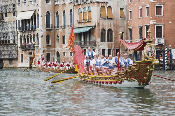 Venice, Veneto, Italy. Participants in the Regata Storico historical regatta held annually in September in red and gold decorated gondola and wearing traditional costume approaching the Rialto bridge with onlookers gathered on the balconies of canalside buildings. Teams represent Sestiere districts of Venice in traditional races. Italy Italia Italian Venice Veneto Venezia Europe European City Regata Regatta Gondola Gondola Gondolas Gondolier Boat Architecture Exterior Water Classic Classical Destination Destinations History Historic Holidaymakers Older Southern Europe Tourism Tourist