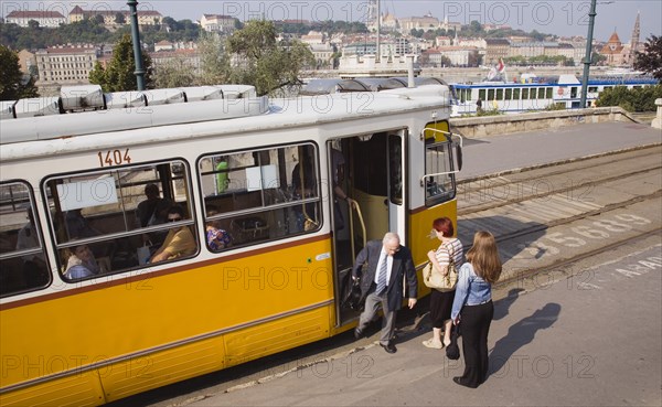 Budapest, Pest County, Hungary. Tram with passengers alighting and view towards the River Danube behind. Hungary Hungarian Europe European East Eastern Buda Pest Budapest City Transport Urban Electric Tram Yellow People Passengers Commuters Commuter Color Destination Destinations Eastern Europe