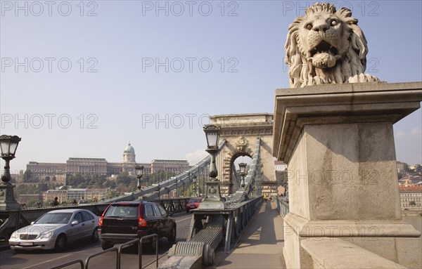 Budapest, Pest County, Hungary. Lion sculpture on the Chain Bridge or Memory Bridge and passing traffic. Hungary Hungarian Europe European East Eastern Buda Pest Budapest City Architecture Transport Chain Memory Bridge Traffic Cars Lion Statue Sculpture Art Automobiles Autos Destination Destinations Eastern Europe