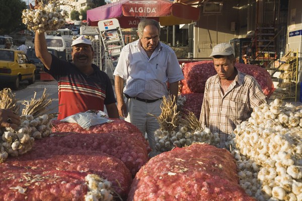 Kusadasi, Aydin Province, Turkey. Stallholder selling garlic at weekly market standing behind stall stacked high with sacks of garlic bulbs and holding up bunch of tied bulbs in late afternoon sunshine. Turkey Turkish Eurasia Eurasian Europe Asia Turkiye Aydin Province Kusadasi Market markets Stall Vendor Man Male Men Garlic Sacks Bulb Bulbs Destination Destinations European Male Man Guy Male Men Guy Middle East South Eastern Europe Western Asia
