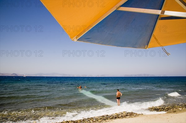 Kos, Dodecanese Islands, Greece. Holidaymakers on beach outside Kos Town with blue and orange striped parasol in foreground and man wearing swimming trunks standing at waters edge in cross made by surf looking towards others in water. Greece Greek Europe European Vacation Holiday Holidays Travel Destination Tourism Ellas Hellenic Docecanese Kos Sea Umbrella Umbrella Parasol Swim Swimmers Man Tourist Color Destination Destinations Ellada Male Men Guy Sand Sandy Beaches Tourism Seaside Shore Tourist Tourists Vacation Sightseeing Southern Europe Sunbather Surfing Surfer Waves Sport Watersport Breaking Breakers Surfboarding Water
