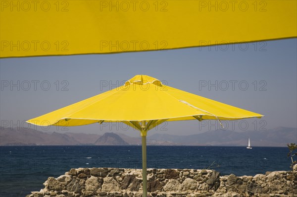 Kos, Dodecanese Islands, Greece. Bright yellow parasols on beach outside Kos Town with crumbling stone wall in foreground and view towards distant coastline beyond. Greece Greek Europe European Vacation Holiday Holidays Travel Destination Tourism Ellas Hellenic Docecanese Kos Sea Umbrella Umbrellas Parasol Parasols Yellow Blue Color Destination Destinations Ellada Sand Sandy Beaches Tourism Seaside Shore Tourist Tourists Vacation Southern Europe Water