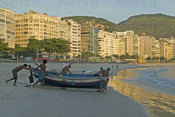 Rio de Janeiro, Brazil. Launching a fishing boat at Copacabana beach at daybreak fishermen and a few early-morning walkers on the beach soft sunlight on the hotel facades. Brazil Brasil Brazilian Brasilian South America Latin Latino American City Beach Fishing Fishermen Fisherman Industry Boat Transport Sea Hotels Architecture. Morning Sunrise Early People Working Destination Destinations Latin America Sand Sandy Beaches Tourism Seaside Shore Tourist Tourists Vacation South America Southern Water