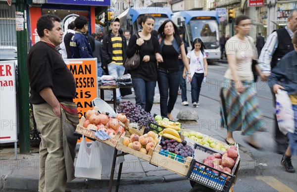 Istanbul, Turkey. Sultanahmet. Pedestrians pass fresh fruit stall with male vendor on busy street beside tramway. Turkey Turkish Istanbul Constantinople Stamboul Stambul City Europe European Asia Asian East West Urban Destination Travel Tourism Sultanahmet Market Market Vendor Vendor Fruit Busy People Crowded Crowd Destination Destinations Middle East South Eastern Europe Turkiye Western Asia