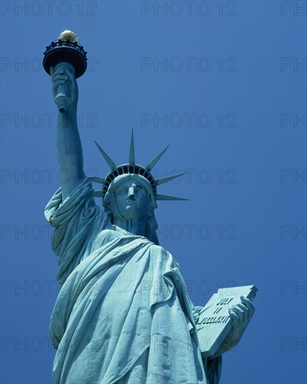 New York, New York State, USA. Statue of Liberty. Detail id upper torso of green statue holding torch with gold flame against a blue sky. American Color Destination Destinations North America Northern United States of America