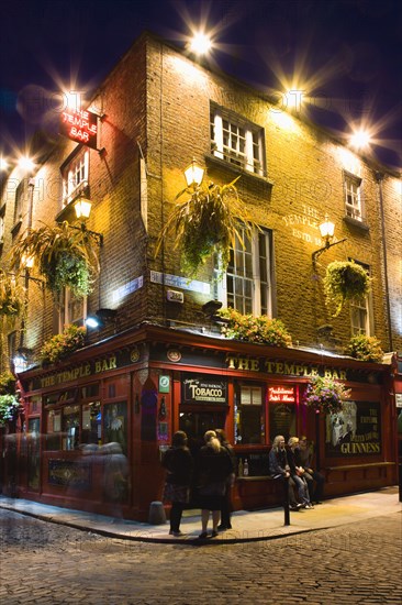 Ireland, County Dublin, Dublin City, Temple Bar Pub illuminated at night with people walking past on the cobbled streets of the area on the south bank of the Liffey River.