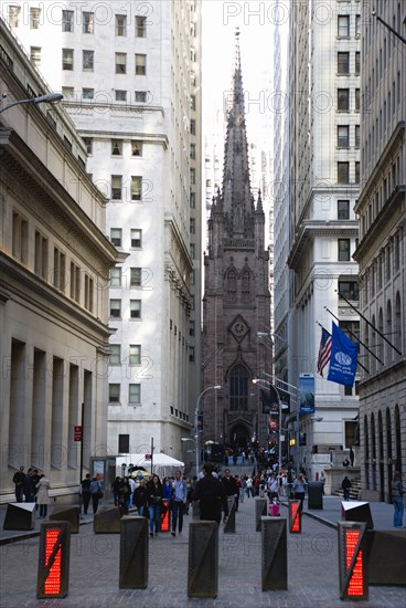 USA, New York, New York City, Manhattan  People walking in Wall Street by security barriers in the road with the Gothic Trinity Church on Broadway at the end of the street.