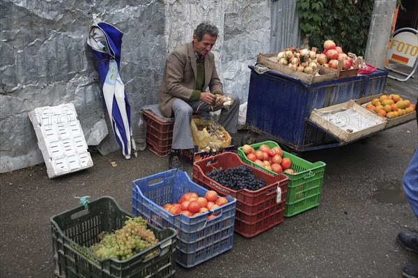 Albania, Tirane, Tirana, Fruit and vegetable street vendor seated on crate to bunch garlic behind display including grapes  oranges and onions. Part seen customer in foreground.