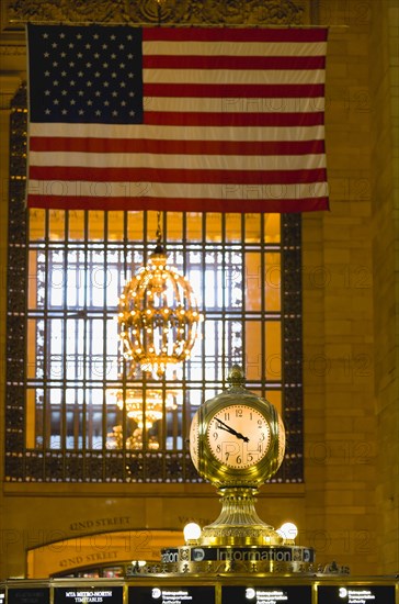 USA, New York, New York City, Manhattan  Grand Central Terminal railway station with the four faced clock above the travel information booth in the Main Concourse beneath a chandelier and the American Stars and Stripes flag hanging from the ceiling.