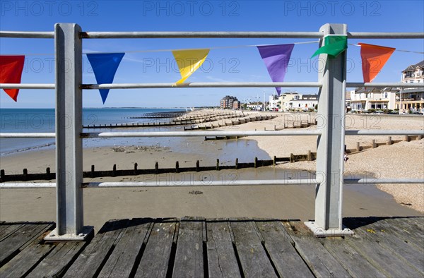 England, West Sussex, Bognor Regis, Wooden groynes at low tide used as sea defences against erosion of the shingle pebble beach seen through metal railings with colourful flags on the pier.