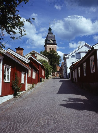Sweden, Sodermanland, Strangnas, Cobbled street lined by wooden houses leading to Domkyrkan Cathedral  part seen exterior facade and clock tower.