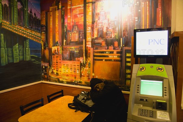 USA, New York, New York City, Manhattan  Man asleep at table beneath wall painting of Manhattan beside an ATM cash machine in Mcdonalds restaurant in Times Square.