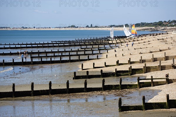 England, West Sussex, Bognor Regis, Wooden groynes at low tide used as sea defences against erosion of the shingle pebble beach with people on the sand by the waterline and small sailing boats on the beach.