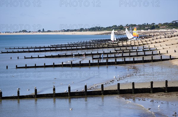 England, West Sussex, Bognor Regis, Wooden groynes at low tide used as sea defences against erosion of the shingle pebble beach with people on the sand by the waterline and small sailing boats on the beach.
