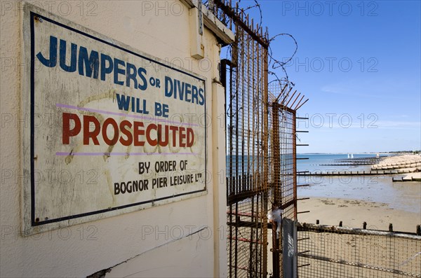 England, West Sussex, Bognor Regis, Warning sign against jumping or diving off the pier into the water on the pier with wooden groynes at low tide used as sea defences against erosion of the shingle pebble beach beyond.