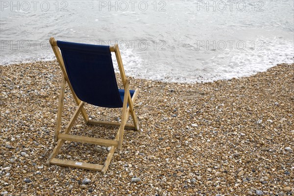 England, West Sussex, Bognor Regis, Single dark blue deck chair on pebble shingle beach looking out to sea with waves gently breaking on the shoreline.