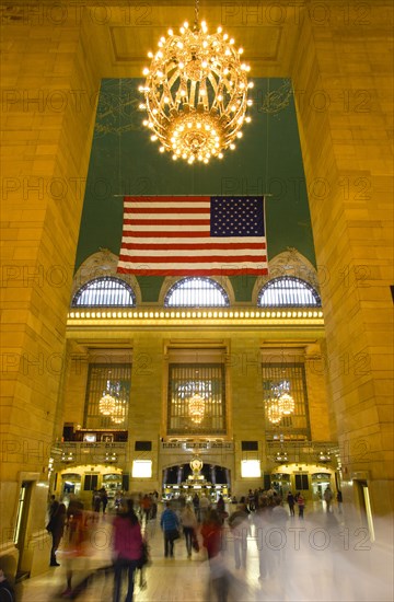USA, New York, New York City, Manhattan  Grand Central Terminal railway station entrance to the Main Concourse from the Vanderbilt Hall with people walking below the American Stars and Stripes flag and chandelier hanging from the ceiling.