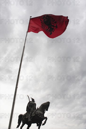 Albania, Tirane, Tirana, Equestrian statue of national hero George Castriot Skanderbeg below national flag depicting black two headed eagle on red background flying against grey sky above.