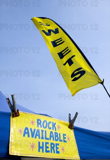 England, West Sussex, Bognor Regis, Yellow flag and sign on a tent that read Sweets and Rock Available Here under a blue sky on the Esplanade beside the beach.