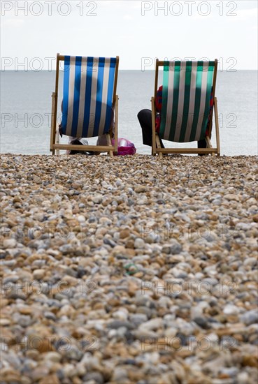 England, West Sussex, Bognor Regis, Two elderly seniors sitting on deck chairs on the pebble shingle beach looking out to sea.