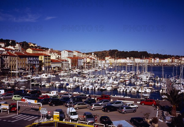 France, Languedoc-Roussillon, Pyrenees Orientales, Port-Vendres.  Inner harbour with moored boats  quay and quayside buildings with car park in foreground. Hillside part visible beyond.
