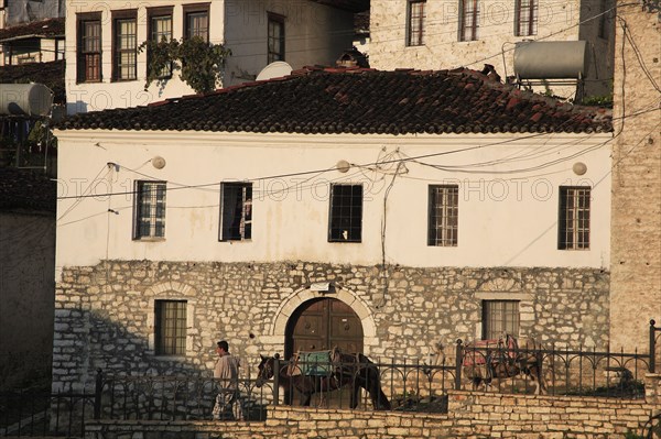 Albania, Berat, Traditional Ottoman buildings with person standing at upstairs window looking down on man leading two horses along street.