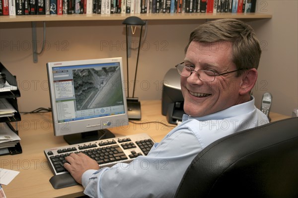 Media, Computer, Office, A man working from his home office  turning towards camera  sitting at desk and using PC. Printer  phone  filing tray of paperwork and shelf of books visible.