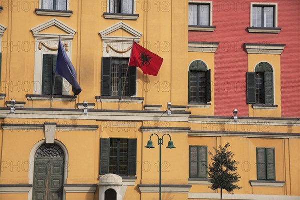 Albania, Tirane, Tirana, Part view of pink and yellow exterior facade of government buildings in Skanderbeg Square flying flags including double headed eagle emblem. Multiple windows with green painted shutters and green painted  arched doorway.