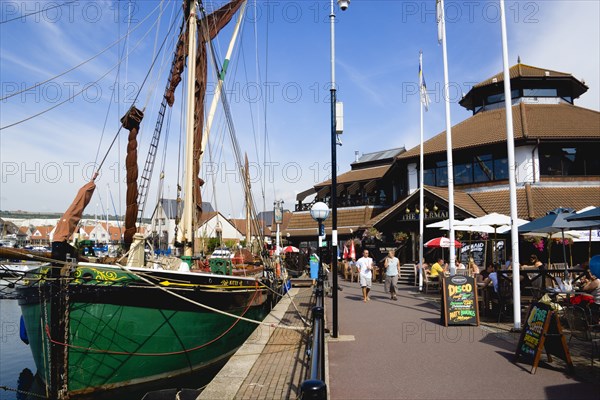 Port Solent Sailing barge SB Kitty moored in the marina with people walking past a pub and restaurant on the dockside and housing beyond. Photo: Paul Seheult