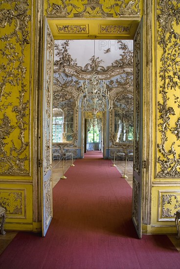 Nymphenburg Palace. Amalienburg The Hall of Mirrors interior of hunting lodge created for Electress Amalia in European Rococo style. Open doorway in yellow painted wall covered with gold decoration and length of red carpet leading through to lavishly d. Photo : Hugh Rooney