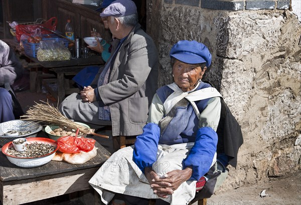 Old Naxi woman wearing traditional costume selling goods on market stall in the village near Lijiang. Photo : Mel Longhurst