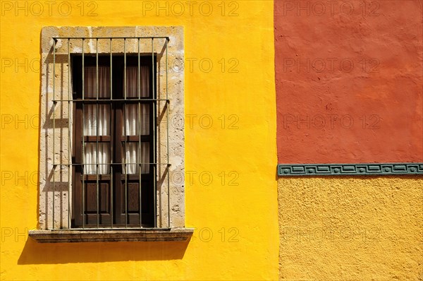 Mexico, Bajio, San Miguel de Allende, Detail of exterior wall of bright yellow and orange painted buildings with window with closed shutter and metal grill. 
Photo : Nick Bonetti