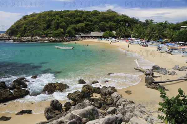 Mexico, Oaxaca, Puerto Escondido, View onto Playa Manzanillo beach with rocks and driftwood in foreground tourist boats people and tree covered headland beyond. 
Photo : Nick Bonetti