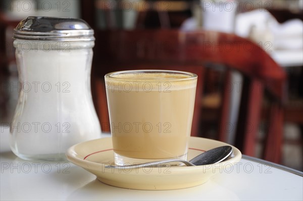 Mexico, Veracruz, Cafe con leche. Coffee with milk served in glass with saucer and spoon on table beside sugar canister. 
Photo : Nick Bonetti