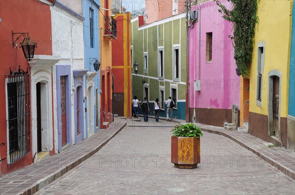 Mexico, Bajio, Guanajuato, Paved street lined by colourfully painted houses group of four people walking at far end and container of geraniums in centre foreground. 
Photo : Nick Bonetti