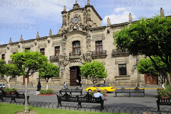 Mexico, Jalisco, Guadalajara, Palacio Gobierno the Government Palace. Exterior facade from Plaza de Armas with passing car trees and man seated on bench in foreground. 
Photo : Nick Bonetti