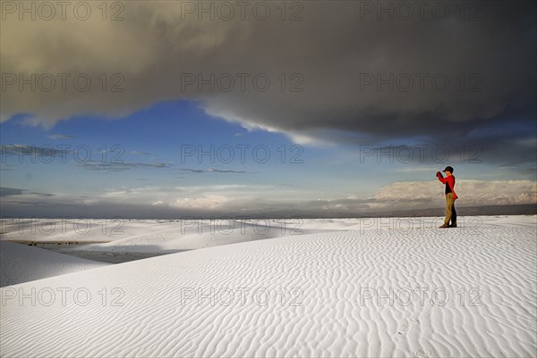 USA, New Mexico, Otero County, White Sands National Monunment. Landscape of white sand with dramatic cloudscape above. Single figure in mid-ground taking photograph. 
Photo : Hugh Rooney